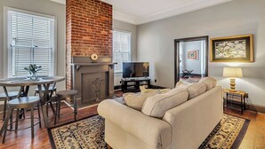 A living room with a brick fireplace, a sofa, a dining table with stools, and a TV. - Historic Garden Flat w/ Screened Porch and Court Yard, Walk to Forsyth & Squares (Savannah)