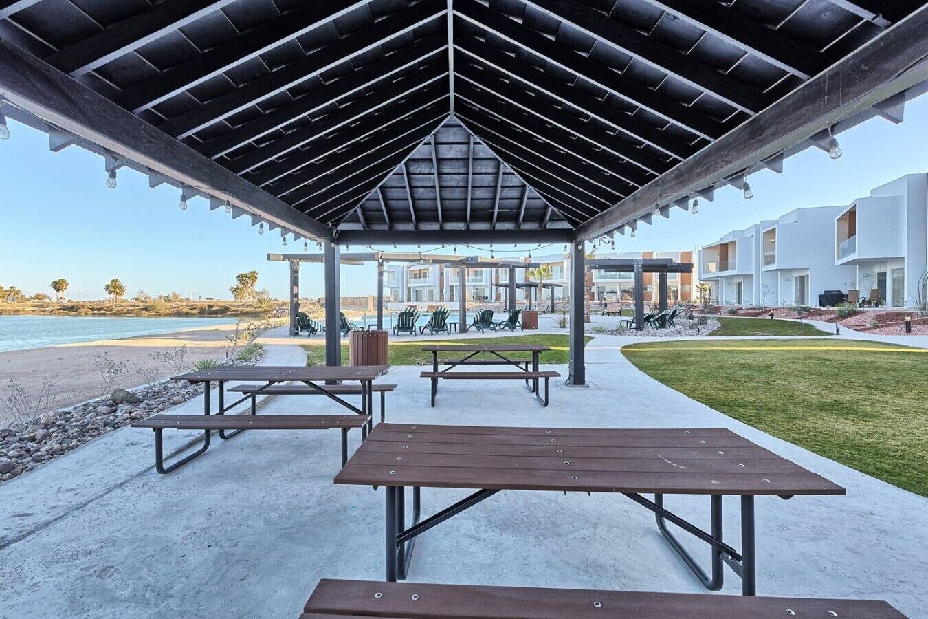 Un espacio al aire libre techado con mesas de picnic, una piscina y edificios modernos al fondo.