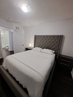 A bedroom with a large bed, a nightstand, a lamp, a door, and a ceiling light.