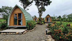 Una fila de pequeñas cabañas de madera con techo verde, rodeadas por un camino de grava y diversas plantas.