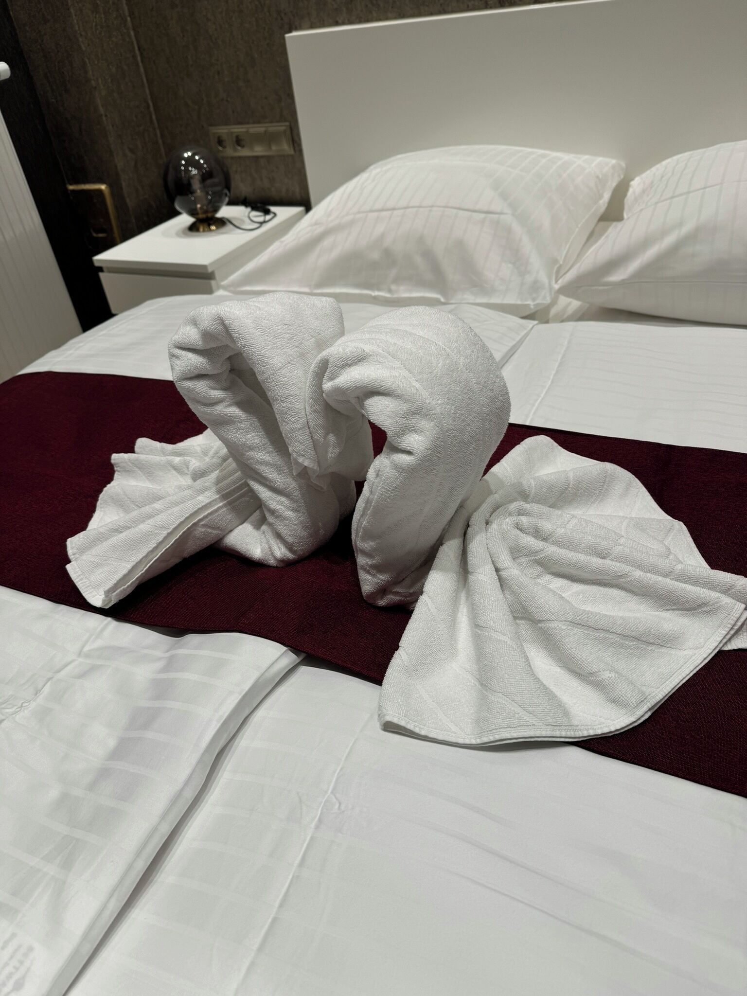 Un lit bien fait, avec du linge de lit blanc et une couverture rouge, une table de chevet avec une lampe et une prise électrique fixée au mur.