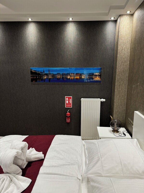 A hotel room with a bed, a radiator, a fire extinguisher, and a cityscape picture on the wall. - Suity-Centrum (Hamburg)