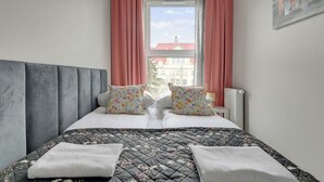 A bedroom with a bed, two pillows, a window with curtains, a radiator, and a painting on the wall. - Mostek 2101 PinkGray by Grand Apartments (Gdańsk)