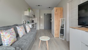 A modern living room with a grey sofa, floral cushions, a small round table, a television, and a kitchen area in the background. - Mostek 2101 PinkGray by Grand Apartments (Gdańsk)