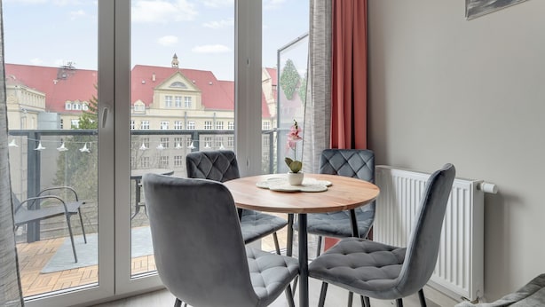 A room with a round table, four grey upholstered chairs, a potted plant, a radiator, and a window with a view of buildings. - Mostek 2101 PinkGray by Grand Apartments (Gdańsk)