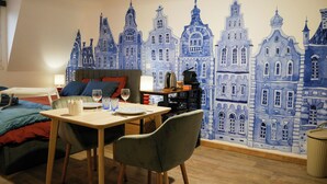 Une pièce comprenant un lit, une table à manger avec des chaises, une lampe et une fresque murale représentant des bâtiments.