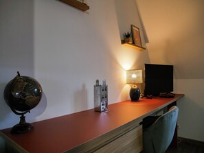 Ein Schreibtisch mit Globus, Lampe und Bildschirm in einem Raum mit schrägem Dach und einem Regal mit einem Bild.