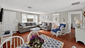 A living room with a grey sofa, white chairs, a small table with a vase of flowers, and a patterned rug. - Waterview Half-Duplex -Walk 2 Seagull Beach/Kayaks (Yarmouth)