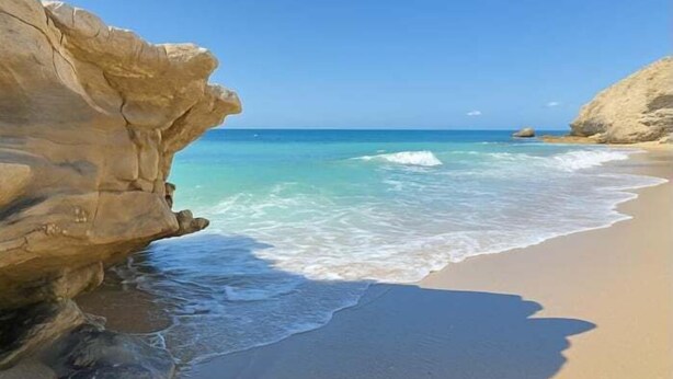 Een strand met helder turquoise water, een grote rotsformatie en een zandstrand.