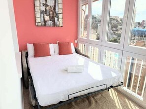 Ein Bett mit weißer Matratze und Kissen, ein Fenster mit Blick auf Gebäude und ein Bild an der Wand.