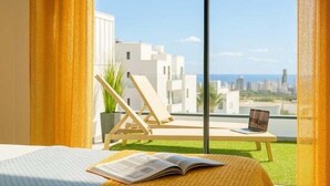 A hotel room with a bed, a window with a view of buildings and the sea, a chair, and a table with a laptop. - Exclusive Apartment. Relax, Luxury and Sea Views (Finestrat)