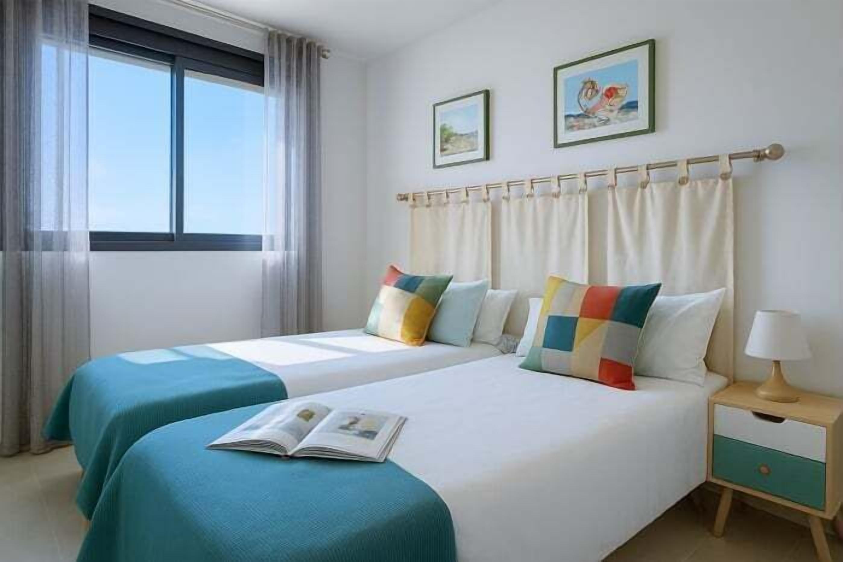 A hotel room with two beds, a window with curtains, a bedside table with a lamp, and two framed pictures on the wall.
