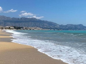 Ein Strand mit klarem Wasser, sandigem Ufer und einer Stadt im Hintergrund.