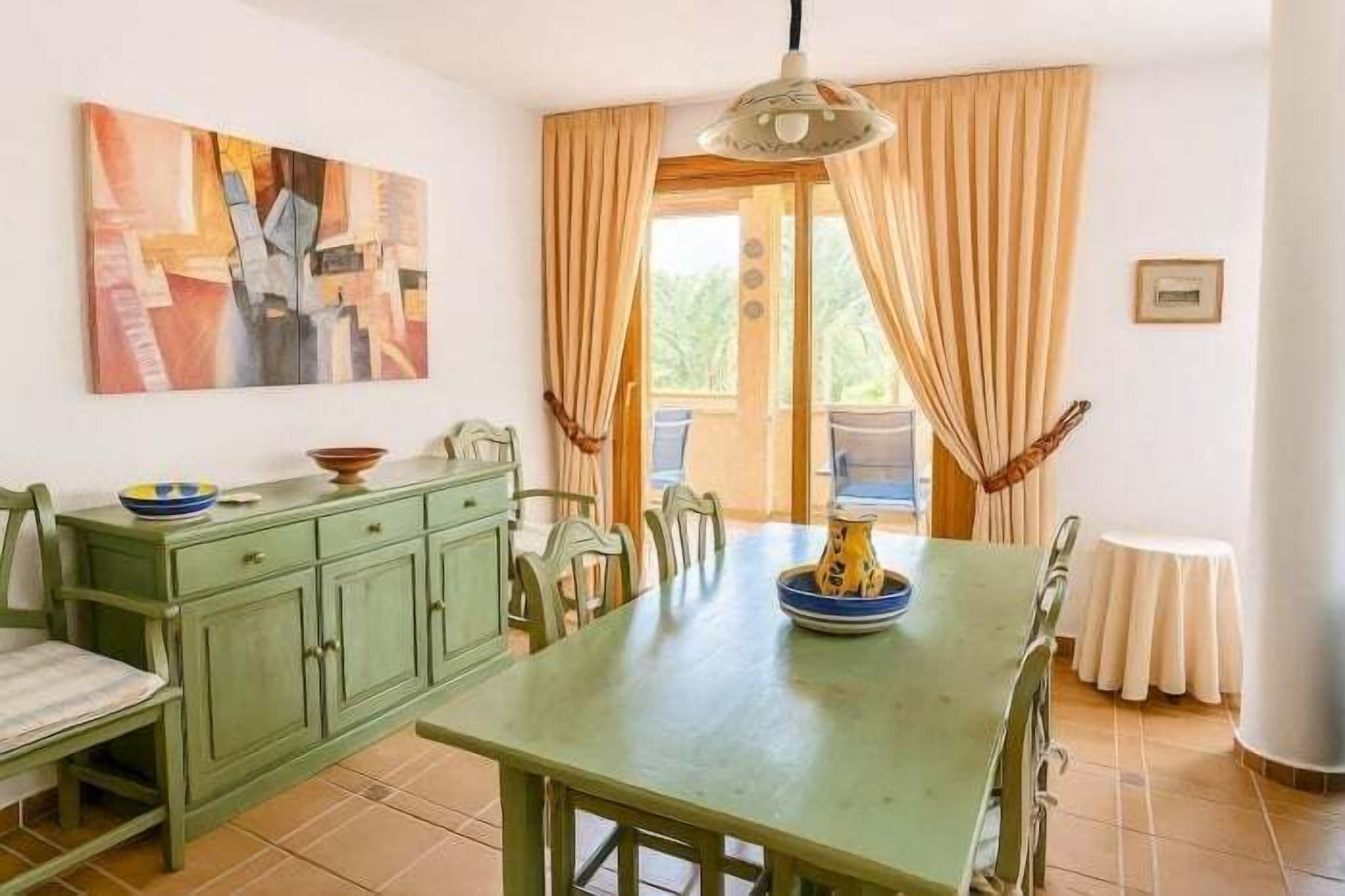 A dining area with a green table, chairs, a painting on the wall, and a view through the window.