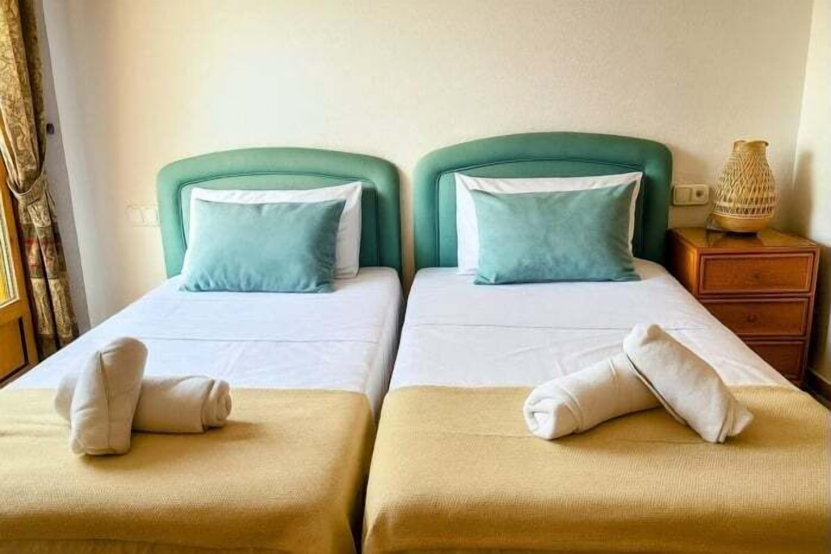 Quarto de hotel com duas camas, cada uma com cabeceira verde, roupa de cama branca e uma pequena mesa de cabeceira de madeira com abajur.