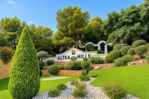 Ein angelegter Gartenbereich mit einem Schild, auf dem 'Altea Hills' steht, umgeben von gepflegtem Grün und einer Steinmauer.