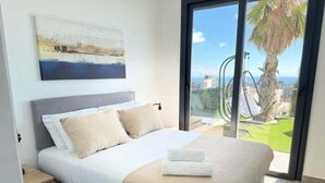 A bedroom with a bed, a nightstand, a painting, and a view of a palm tree and a fence. - Beautiful villa with private pool and sea views (Polop)