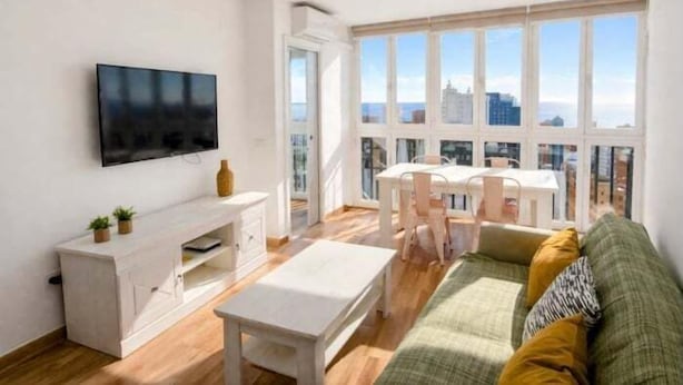 Un salon moderne doté d’un téléviseur à écran plat, d’un meuble de rangement blanc, d’un canapé, d’une table à manger avec des chaises et de grandes fenêtres offrant une vue sur la ville.