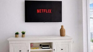 Un téléviseur fixé au mur, orné du logo Netflix, un meuble blanc en dessous avec un vase et deux petites plantes, et une porte avec un trou de serrure.