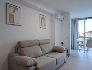 Ein modernes Wohnzimmer mit einem beigen Sofa, zwei gerahmten abstrakten Kunstwerken, einer Klimaanlage und einem Balkon mit Blick.