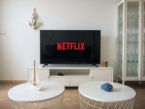 Ein Wohnzimmer mit einem Flachbildfernseher, der 'NETFLIX' anzeigt, zwei runden Tischen, einem dekorativen Segelboot und einer Vitrine mit Glastüren.