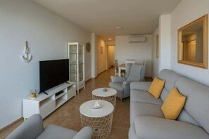 Ein modernes Wohnzimmer mit einem Flachbildfernseher, grauen Sofas und einem Essbereich im Hintergrund.