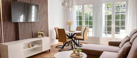 Una sala de estar moderna con un televisor de pantalla plana, un sofá, una mesa de comedor con sillas y un amplio ventanal con vistas.