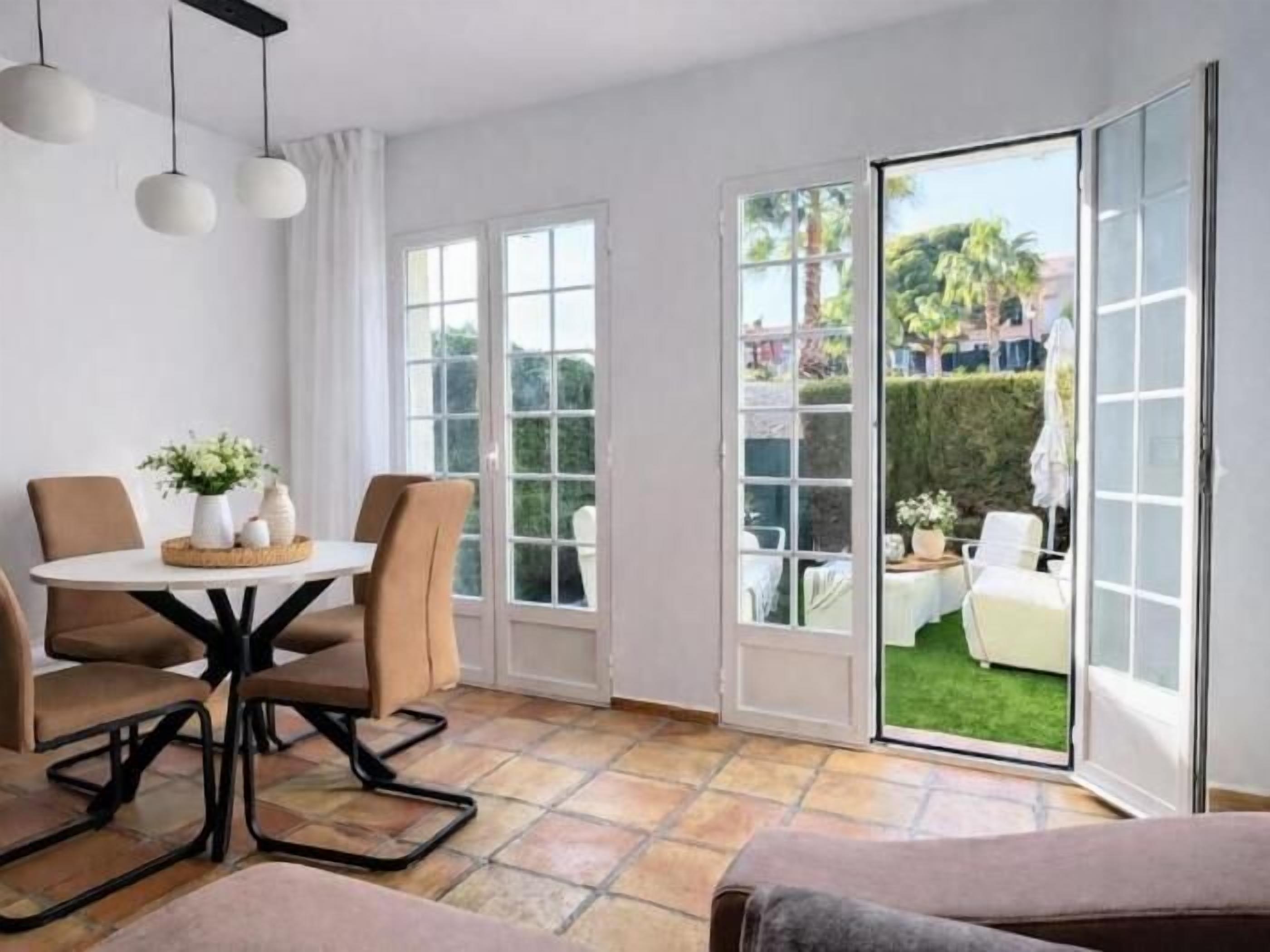 Un espace repas comprenant une table et des chaises, une vue sur le jardin à travers des portes vitrées et un plafond orné de luminaires suspendus.