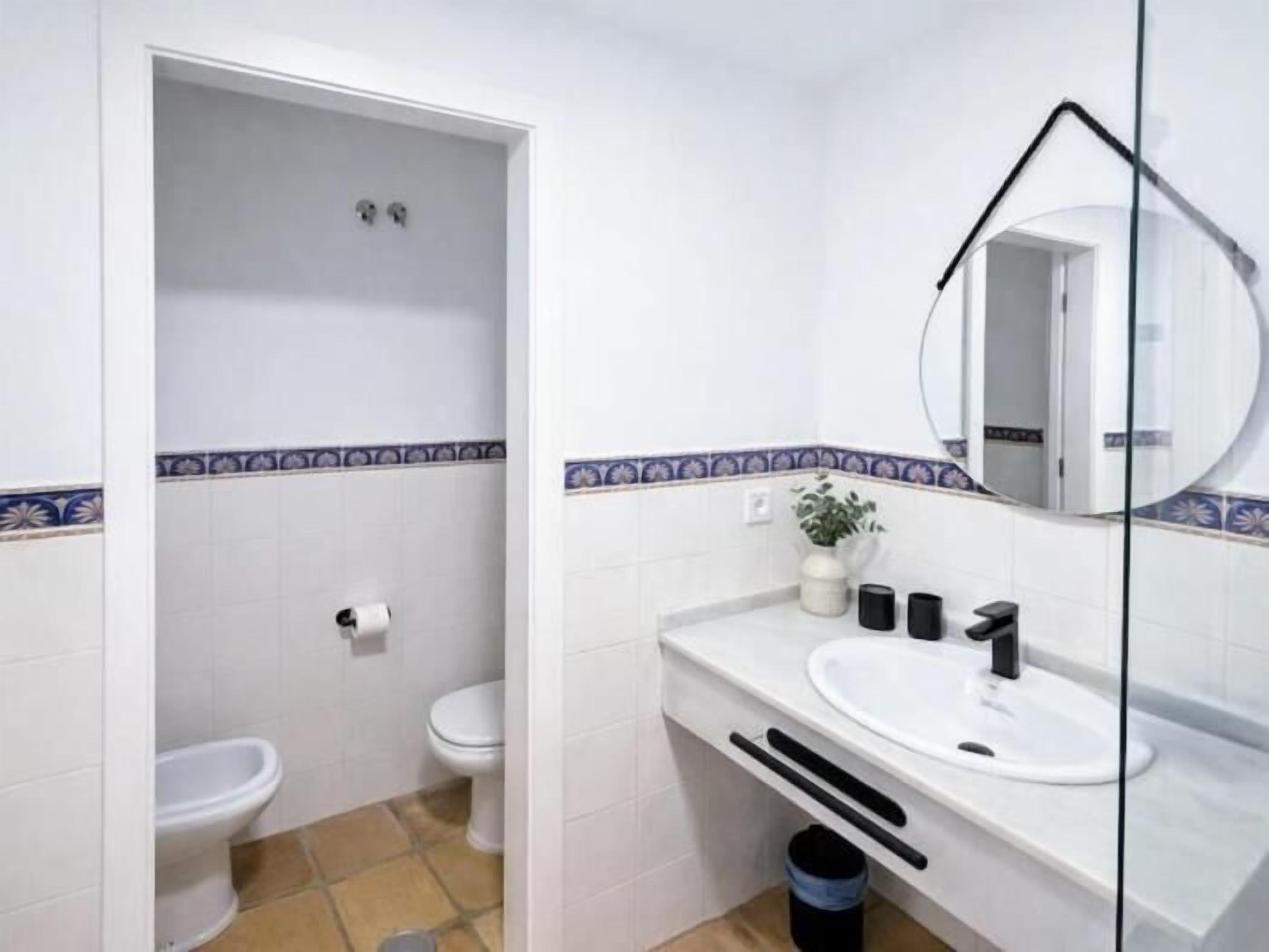 A bathroom with a toilet, bidet, sink, mirror, and a trash bin.