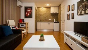 Ein modernes Wohnzimmer mit einer Couch, einem Couchtisch mit Pflanze, einem Fernseher und einer Küchenzeile mit Schränken und einer Mikrowelle.