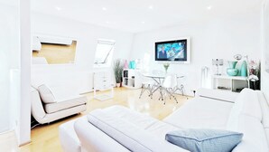 Ein modernes, helles Wohnzimmer mit einem weißen Sofa, einem Essbereich mit einem weißen Tisch und Stühlen sowie einem an der Wand montierten Flachbildfernseher.