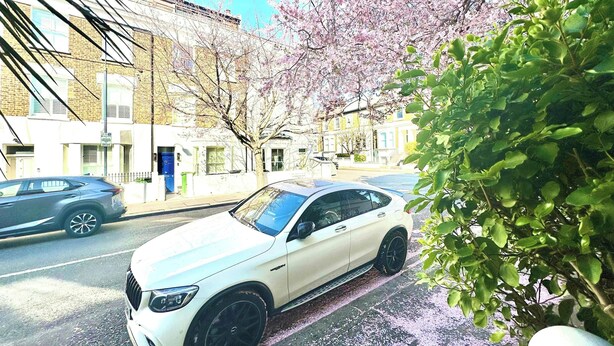 Ein weißes Auto steht auf einer Straße, im Hintergrund sind Gebäude und ein Baum zu sehen.