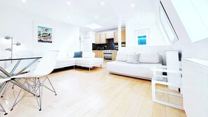 Ein modernes Wohnzimmer mit einem Glastisch, weißen Sofas und einer Küche im Hintergrund.