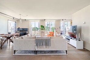 Ein modernes Wohnzimmer mit Sofa, Fernseher, Essbereich und großen Fenstern.