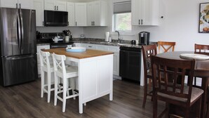 Una cocina moderna con armarios blancos, electrodomésticos de acero inoxidable, una mesa de comedor de madera y un ventanal con persianas.