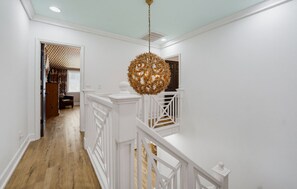 Un couloir doté d’un plancher en bois, de murs blancs et d’une rambarde blanche. Un luminaire décoratif de forme sphérique est suspendu au plafond.