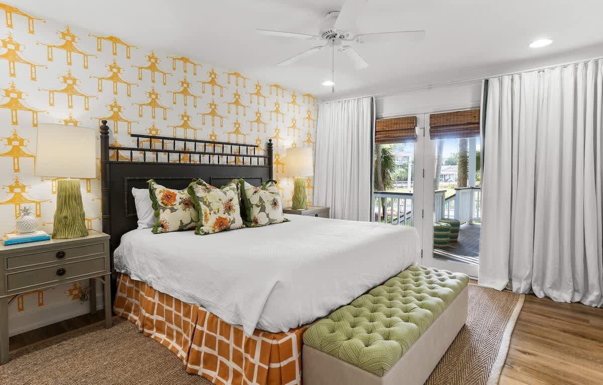 Quarto com cama, mesas de cabeceira, um banco, ventoinha no teto e vista através de uma porta de correr.