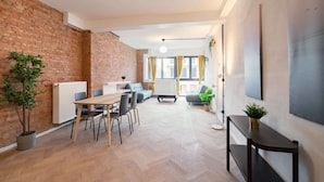 Un espacio habitable moderno con pared de ladrillo, suelo de madera, zona de comedor con sillas, sofá y un amplio ventanal con cortinas.