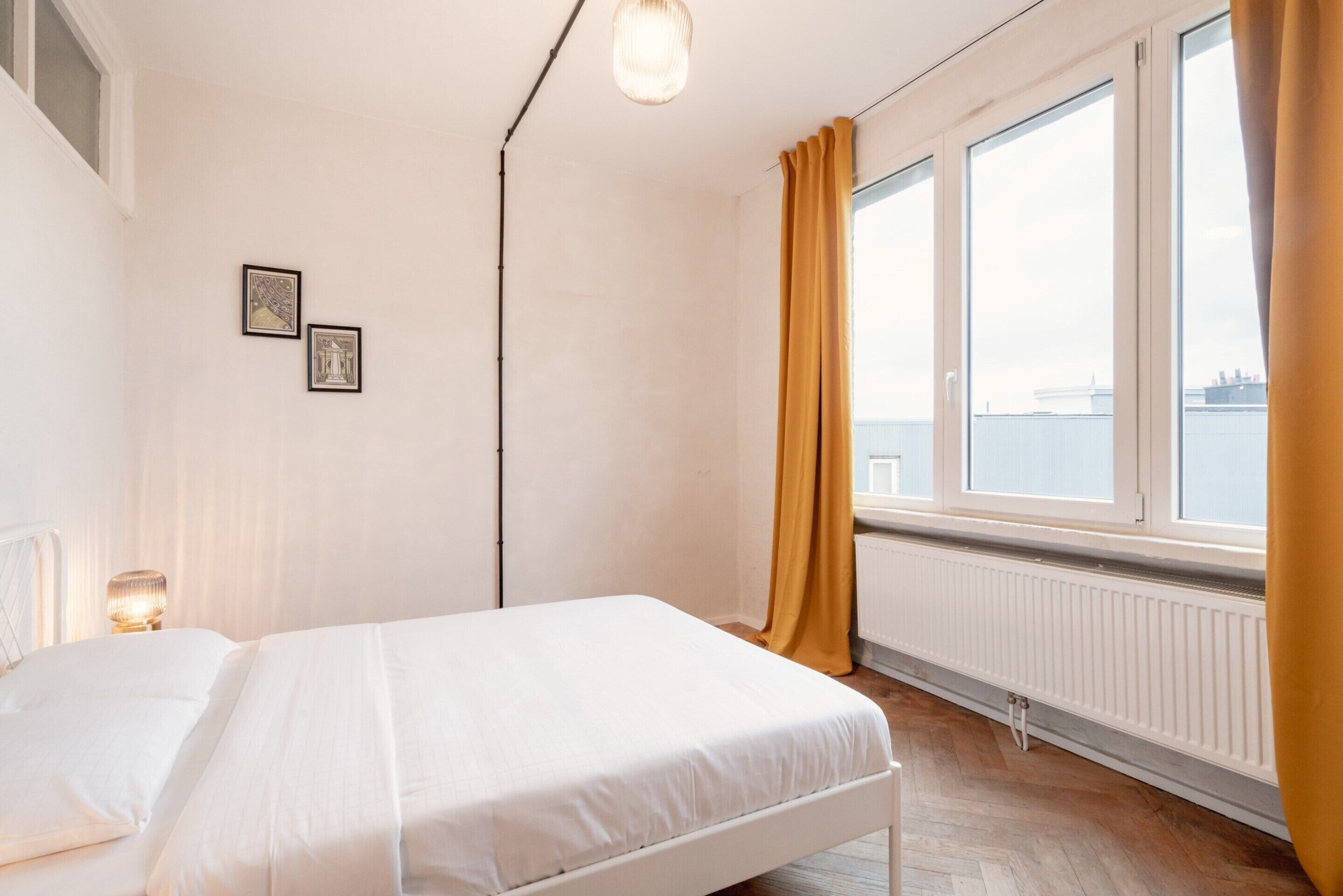 A bedroom with a bed, a window with curtains, a radiator, and two framed pictures on the wall.