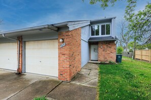 A residential garage with a brick facade, a white garage door, and a small front yard with a wooden fence and a trash bin. - 3-bed/2-bath Townhouse, Near Hospital & Parks (Troy)