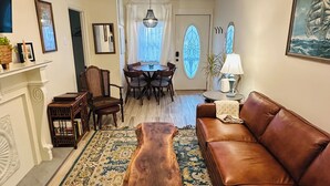 A living room with a brown leather sofa, a wooden coffee table, a patterned rug, a dining area with a chandelier, and a painting of a ship on the wall.