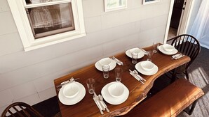 A dining table set with white plates, glasses, and cutlery, flanked by dark wooden chairs, against a light-colored wall with framed artwork.