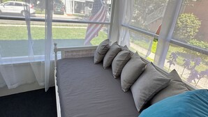 A room with a bed, a window with sheer curtains, an American flag, and a view of a residential street.