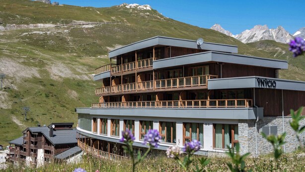 Un hotel de varios pisos con balcones, un telesilla al fondo y un paisaje montañoso.
