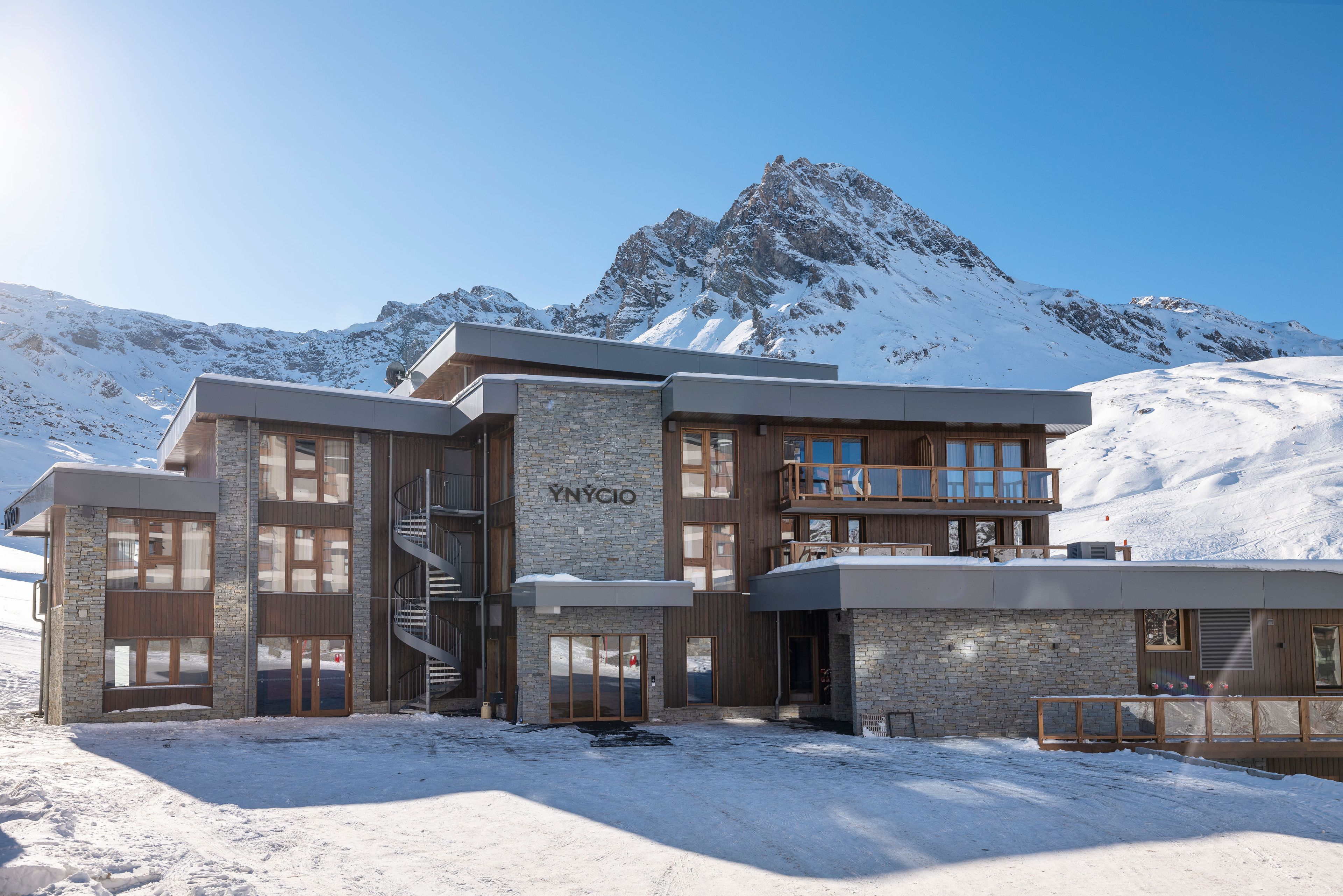 Ein modernes Hotel mit Stein-Fassade, mehreren Balkonen und Blick auf schneebedeckte Berge.