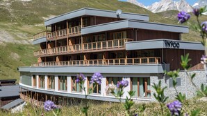 Un hotel de varios pisos con balcones, fachada de piedra y montañas al fondo.