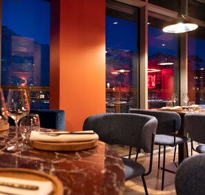A restaurant interior with a dining table set for two, chairs, and a view of the cityscape through large windows.