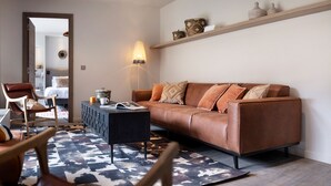 Una sala de estar con un sofá de cuero marrón, un tapete estampado, una mesa de centro negra, una mesita de noche de madera y una estantería en la pared con objetos decorativos.
