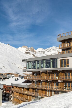A multi-story building with a modern design, featuring balconies and large windows, set against a snowy mountain backdrop.