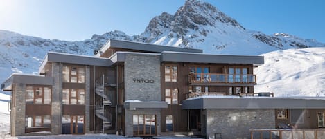 A modern hotel with a stone facade, multiple balconies, and a view of snow-covered mountains.